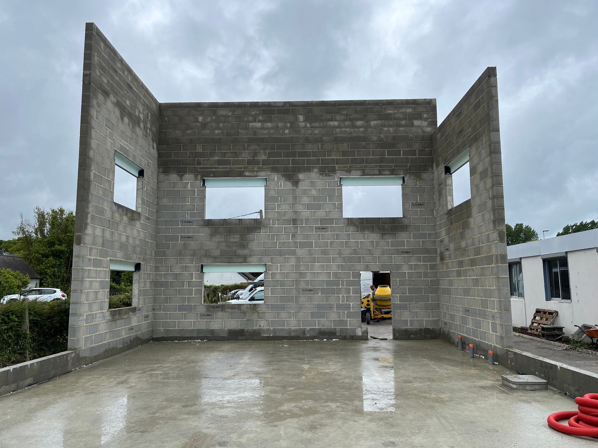 Structure en parpaings partiellement construite, avec ouvertures pour fenêtres et portes, sur une dalle de béton humide.