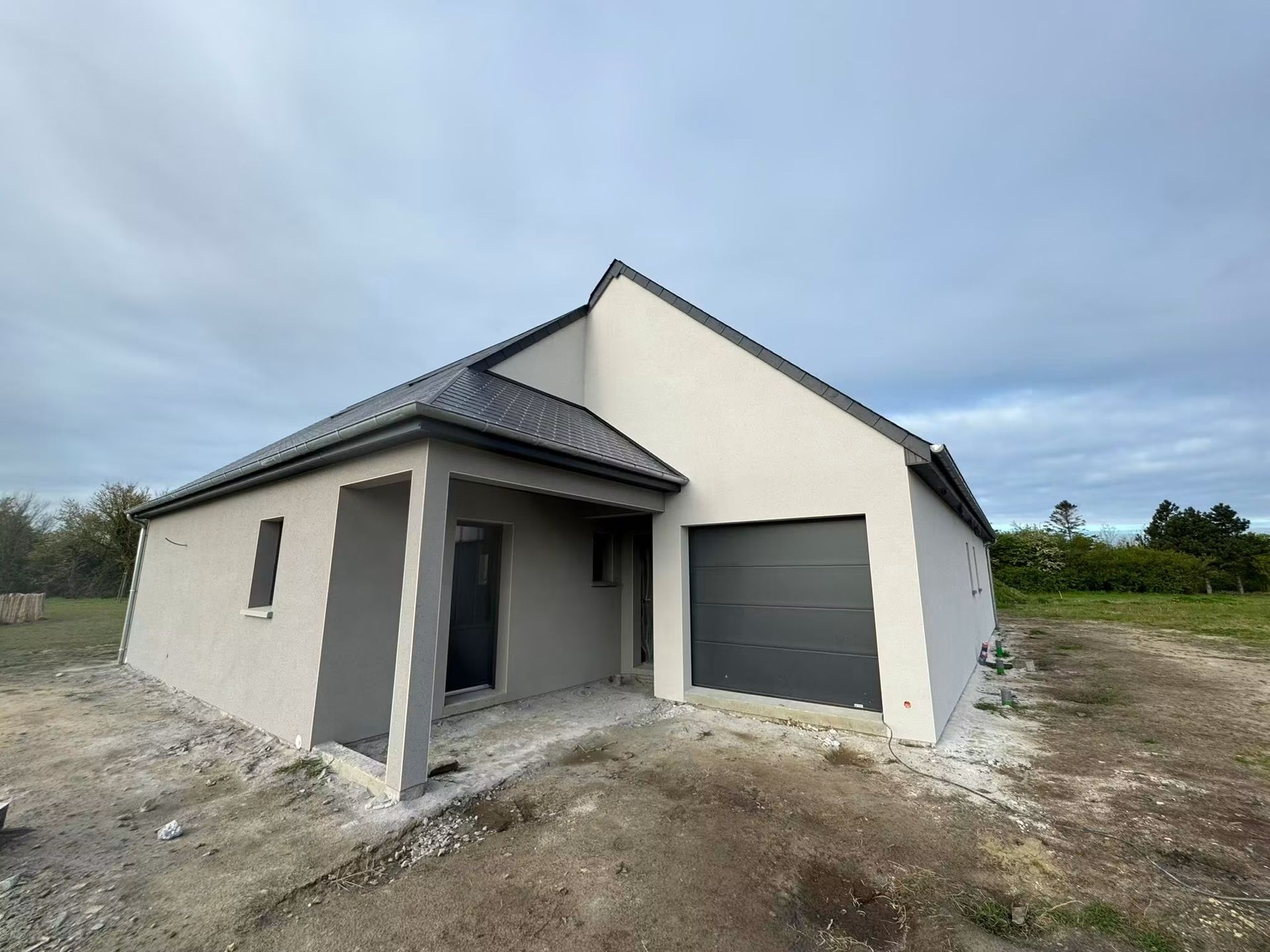 Maison moderne de plain-pied en construction, avec murs beiges, toit gris et cour en terre battue non aménagée.