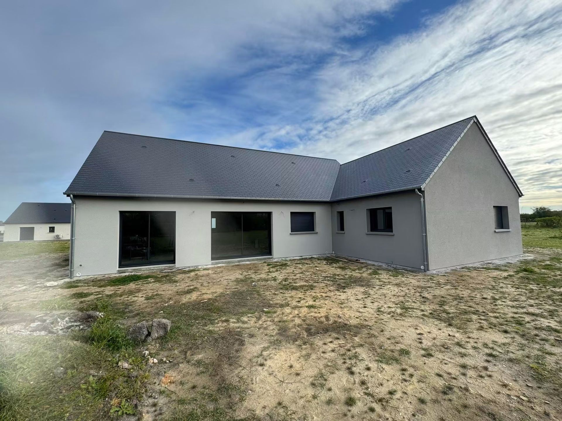Maison blanche neuve avec toit gris et patio couvert sur un terrain sablonneux sous un ciel nuageux