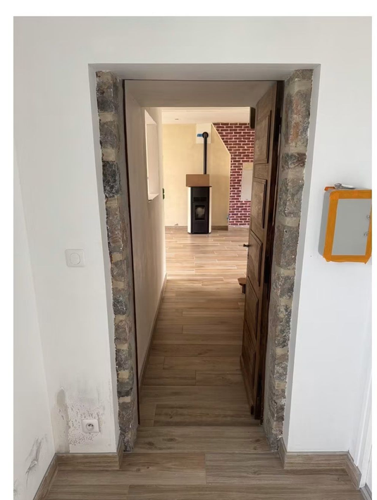 Porte étroite donnant sur une pièce avec parquet, éléments en brique et poêle à bois.