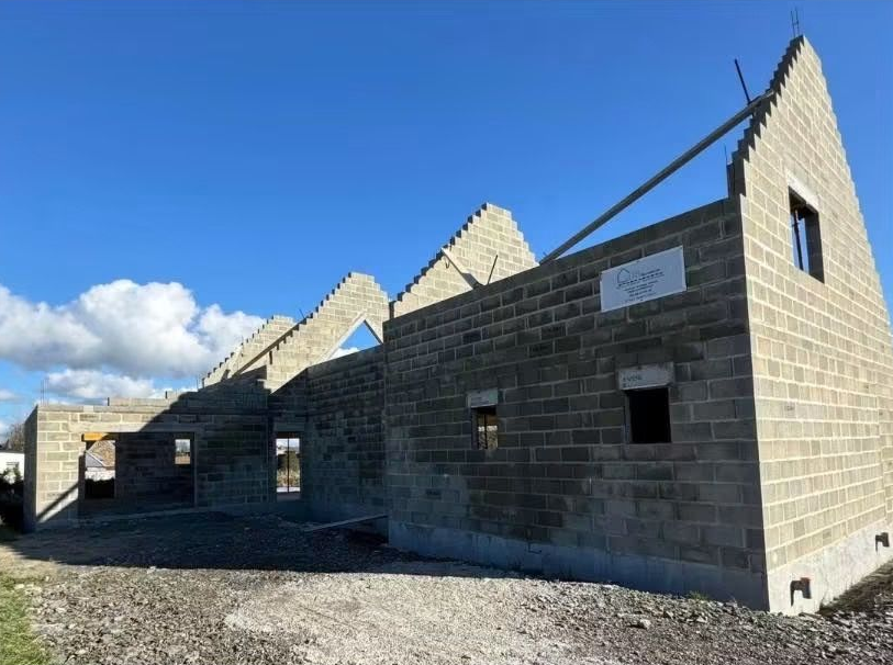 Bâtiment en parpaings partiellement construit, avec une charpente à pignons, sous un ciel bleu.