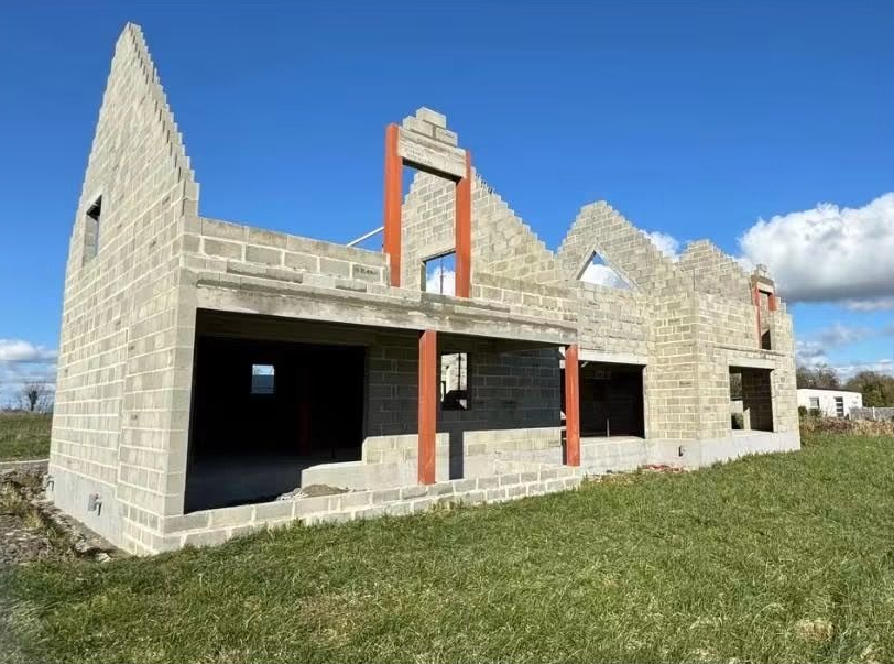 Maison en pierre en construction, murs inachevés, poutres de soutien orange et premier plan herbeux sous un ciel bleu