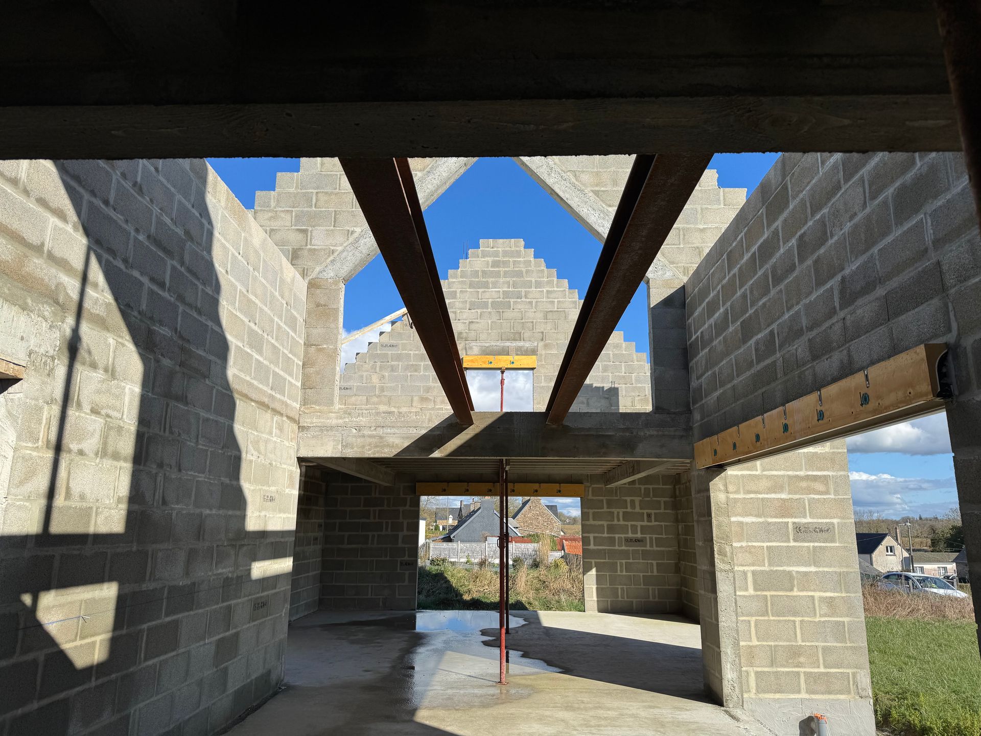 Intérieur d'un bâtiment en parpaings en construction, avec poutres de toit apparentes et fenêtres ouvertes sur le ciel bleu.