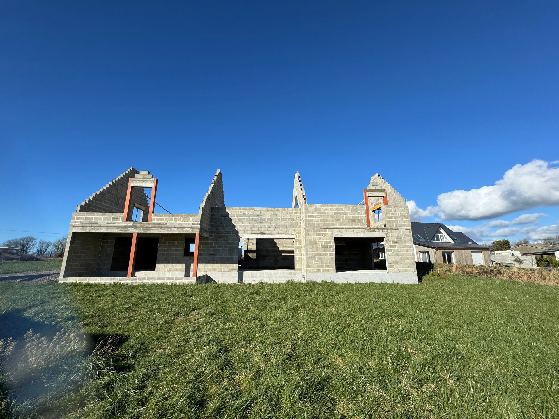 Maison en béton partiellement construite sous un ciel bleu, avec un premier plan herbeux et un littoral rocheux.