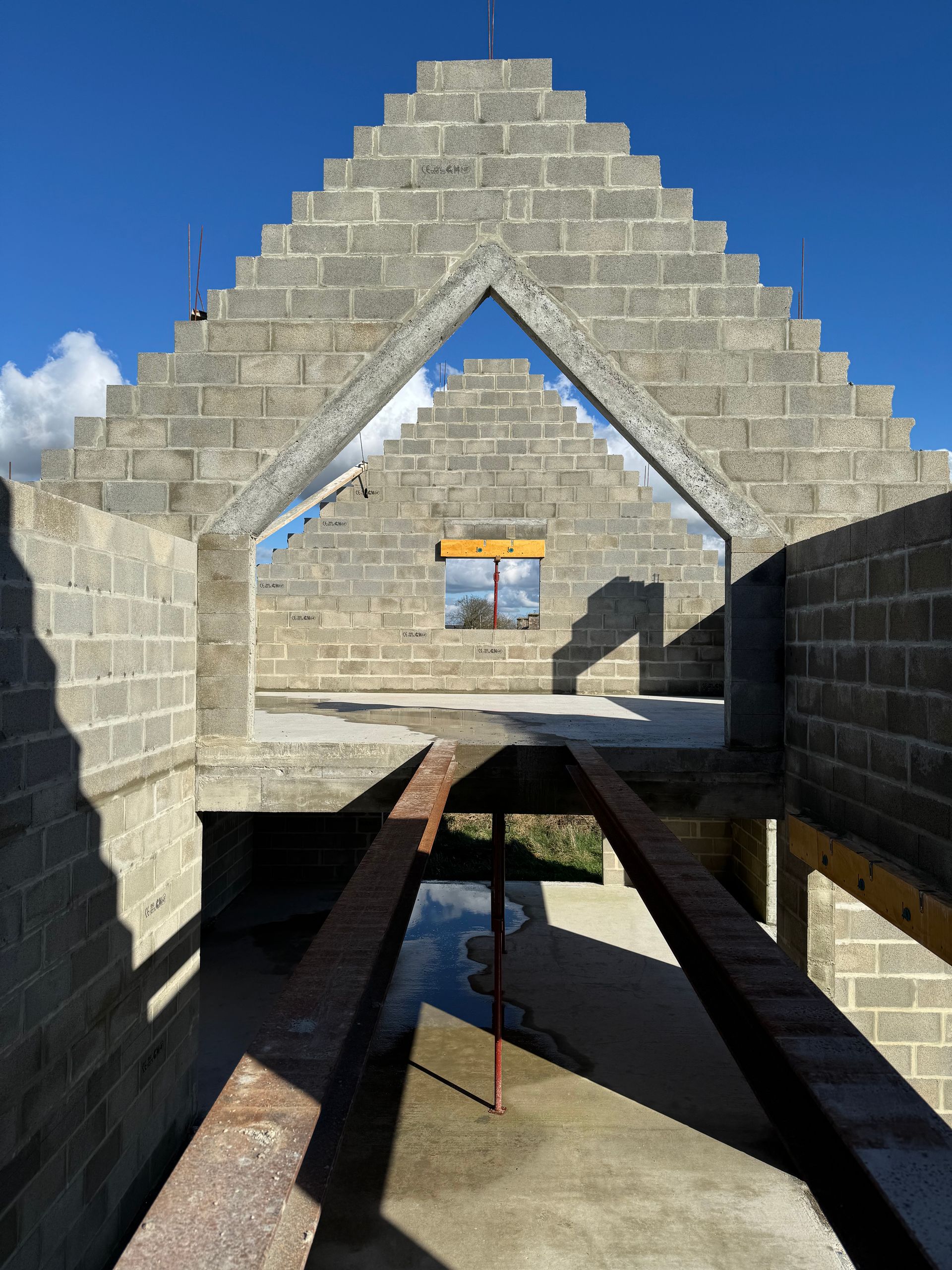 Bâtiment en pierre en construction, avec des murs triangulaires et une allée centrale, sous un ciel d'un bleu éclatant.