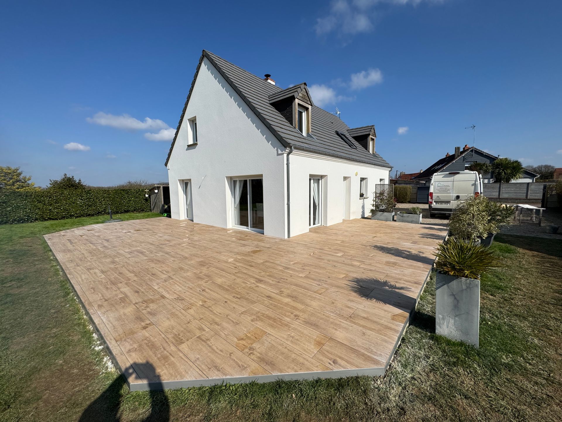 Maison blanche avec toit pentu et grande terrasse pavée dans un jardin ensoleillé