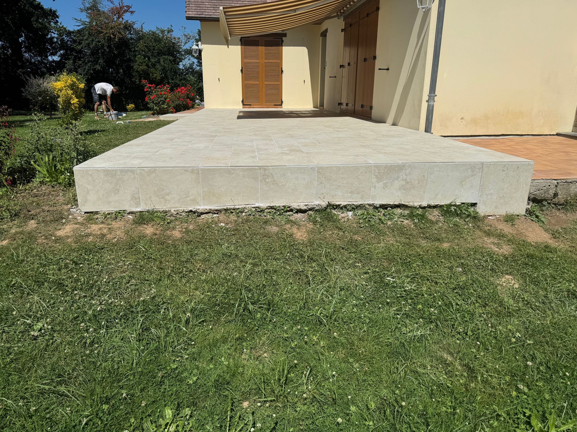 Dalle de terrasse en béton à côté d'une maison, avec de l'herbe devant et une personne travaillant dans le jardin en arrière-plan.