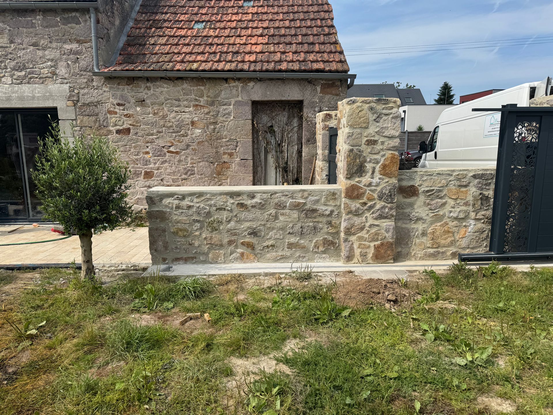 Maison en pierre avec un muret et un portail, donnant sur une cour herbeuse sous un toit de tuiles.
