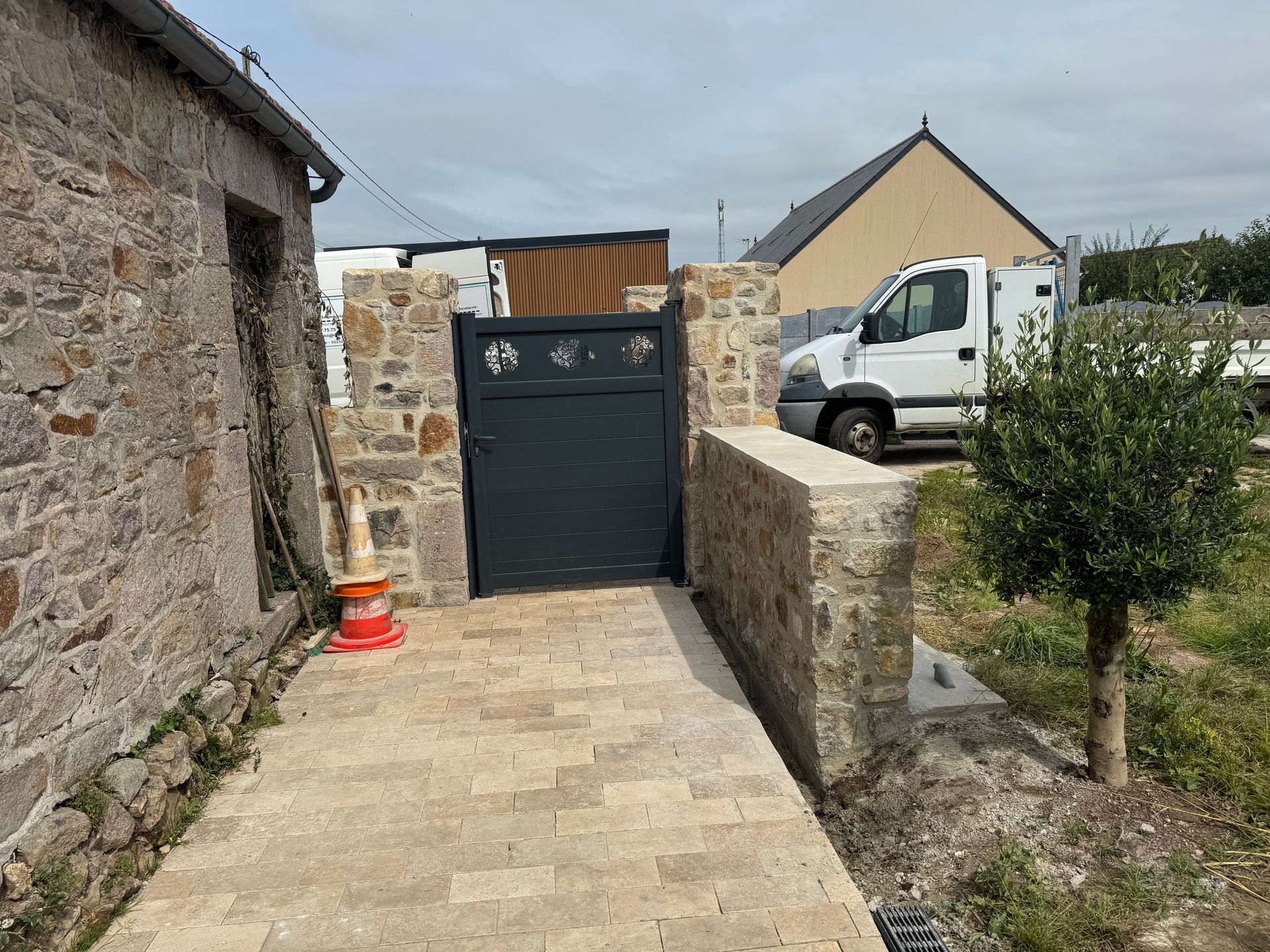 Allée en pierre longeant le mur et le portail, avec une camionnette blanche garée au-delà.