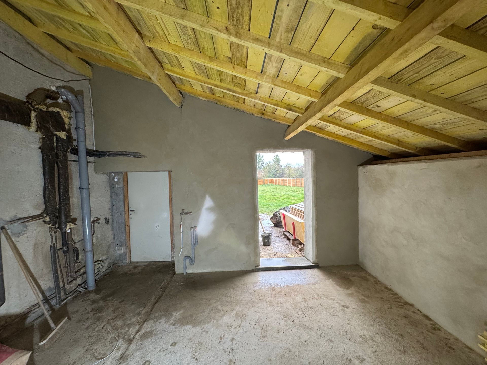 Pièce inachevée avec toit en bois apparent, murs gris et porte ouverte donnant sur une cour gazonnée.