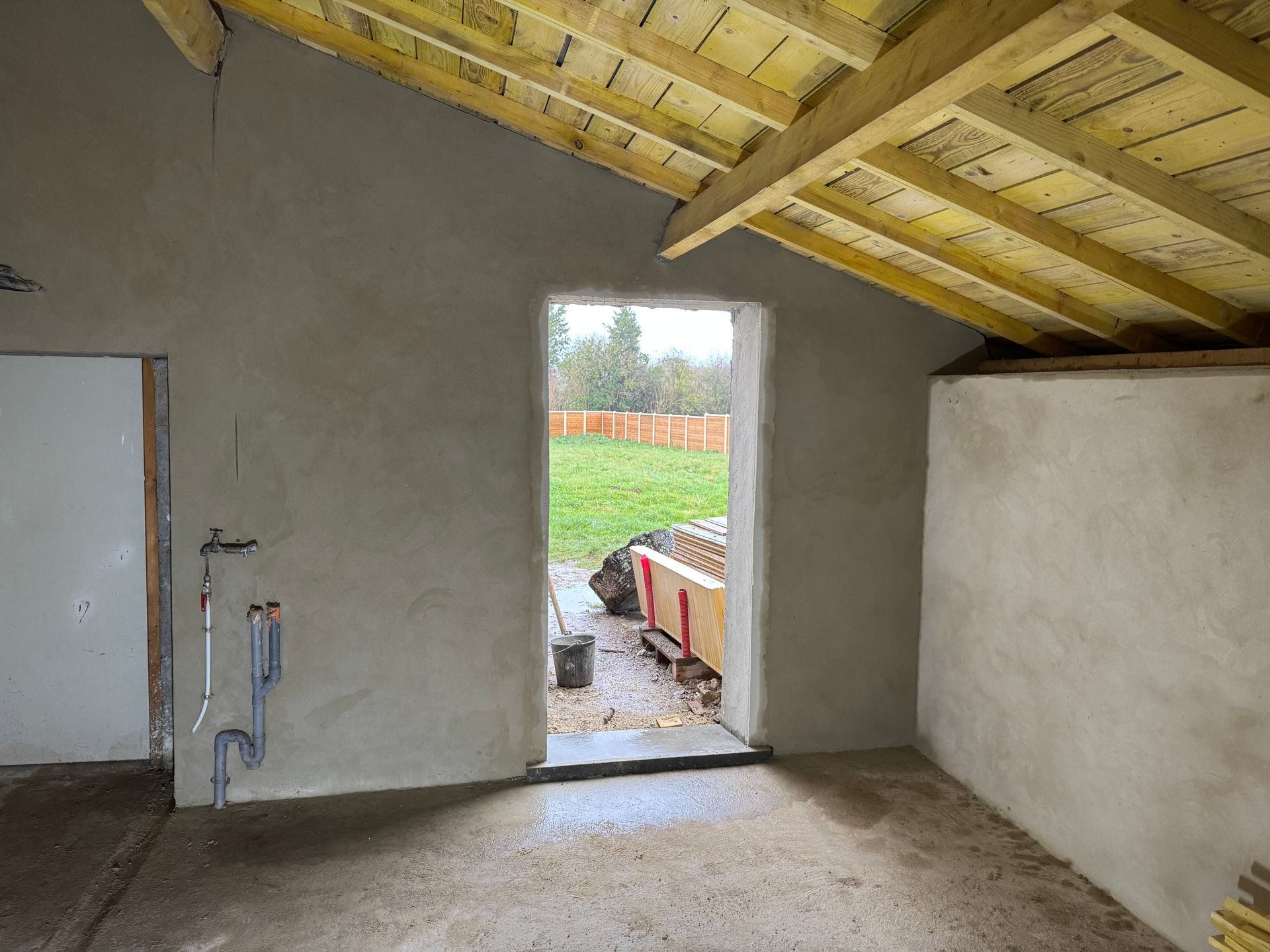 Pièce inachevée avec murs plâtrés, plafond en bois apparent et porte ouverte donnant sur une cour gazonnée.