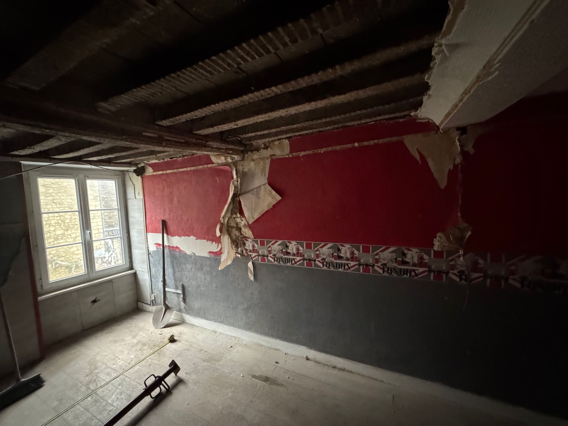 Pièce endommagée avec des murs rouges et gris déchirés, des poutres de plafond apparentes et des débris au sol près d'une fenêtre.