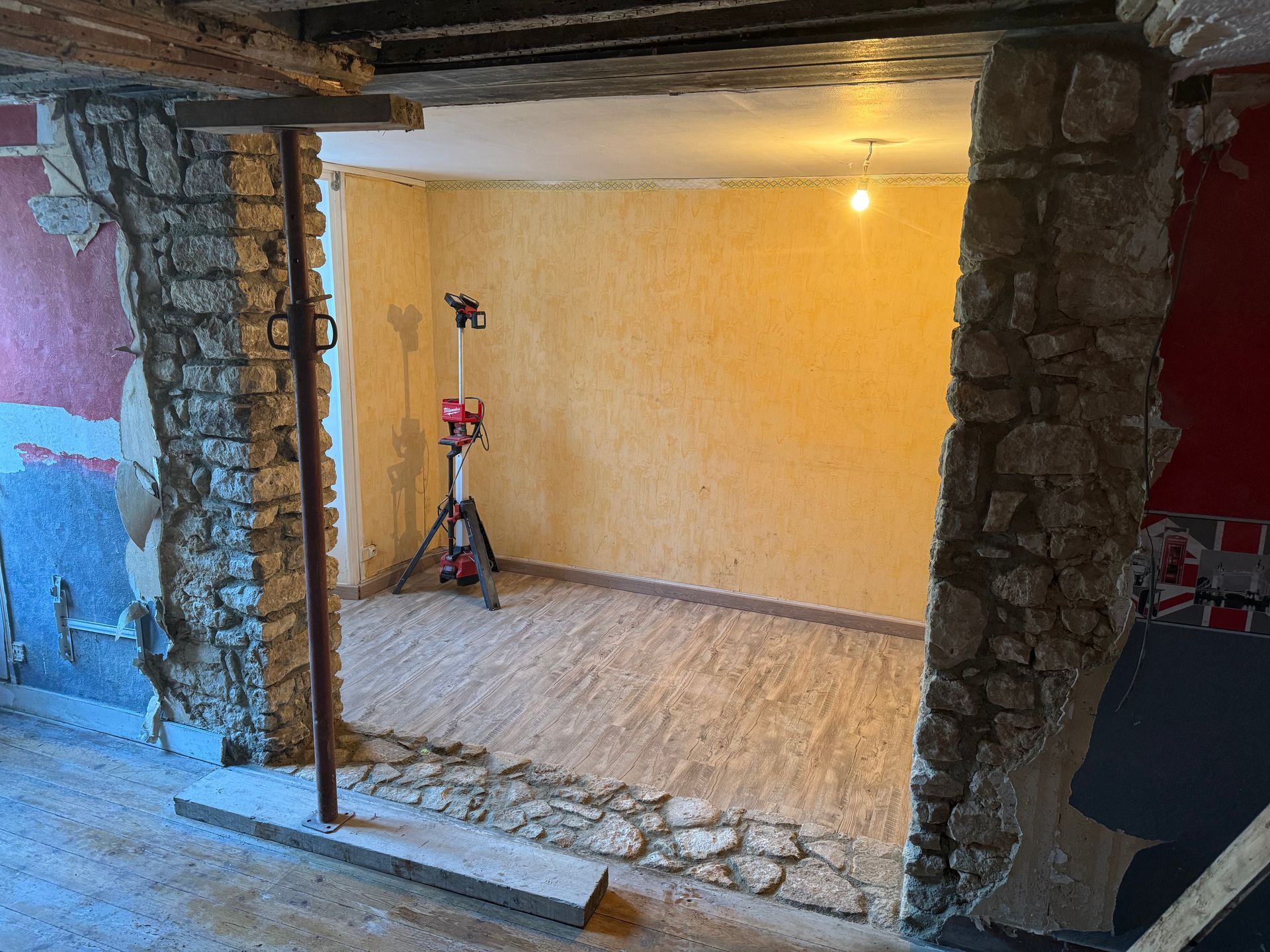 Une porte encadrée de pierre s'ouvre sur une pièce éclairée et vide, aux murs orange, avec un trépied à l'intérieur.