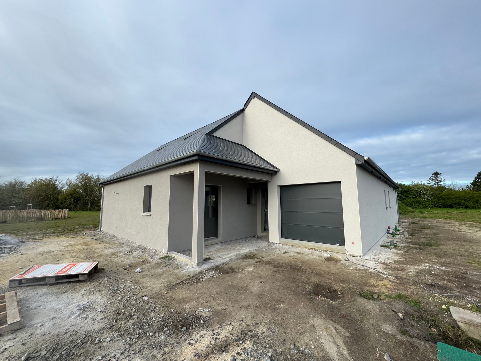 Maison neuve de plain-pied avec toit gris, murs blancs et porte de garage fermée sur un terrain nu