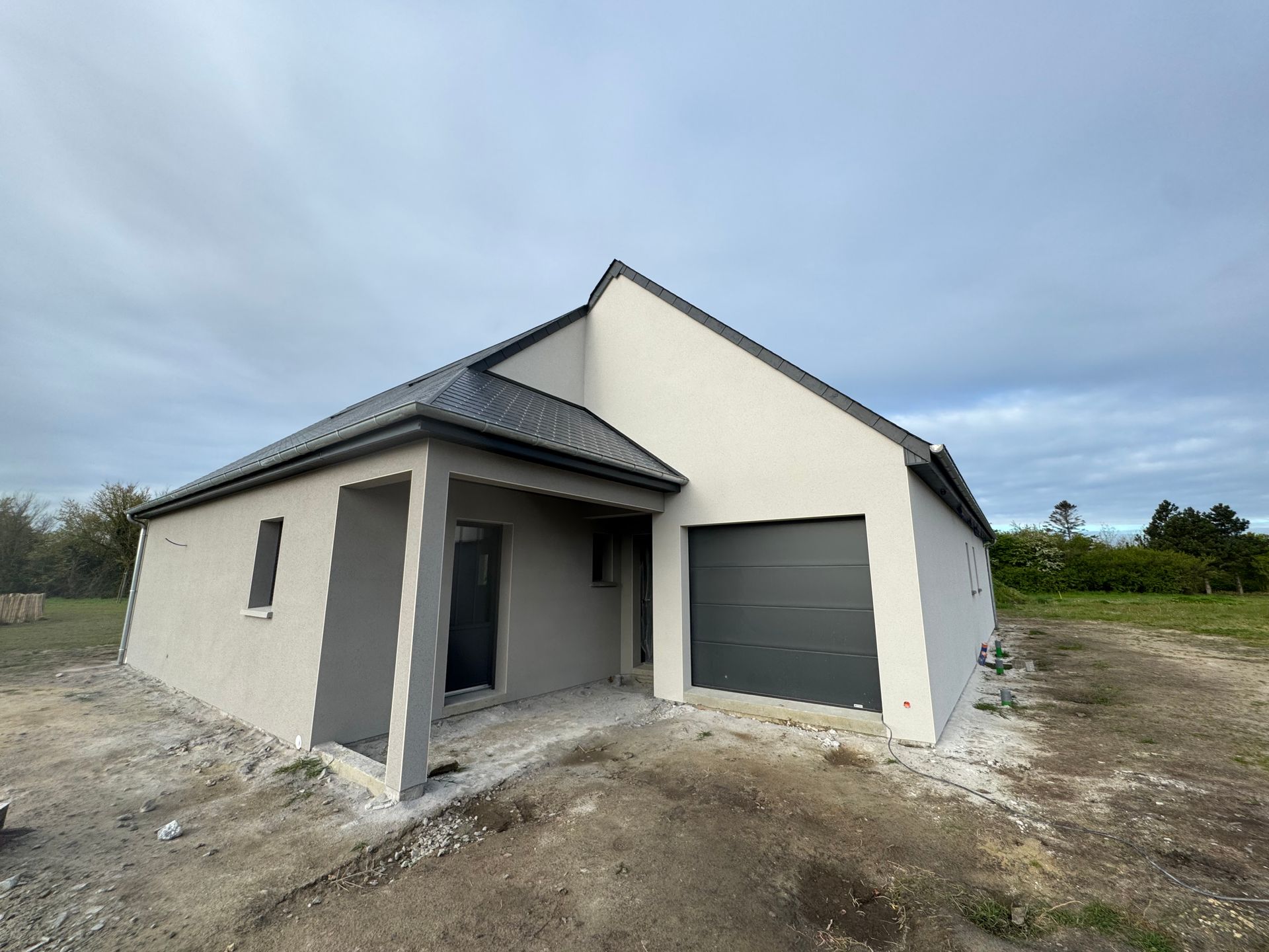 Maison moderne neuve avec porte de garage grise et cour avant en terre battue sous un ciel nuageux