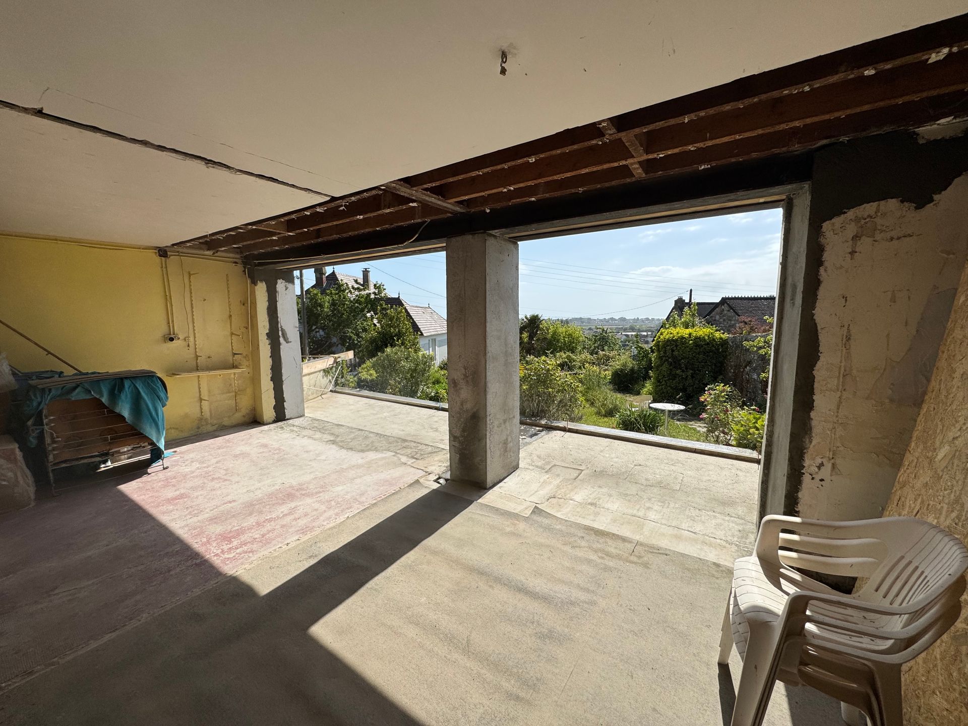 Pièce ouverte avec sol en béton, chaise blanche et vue dégagée sur un paysage verdoyant et des maisons au loin.