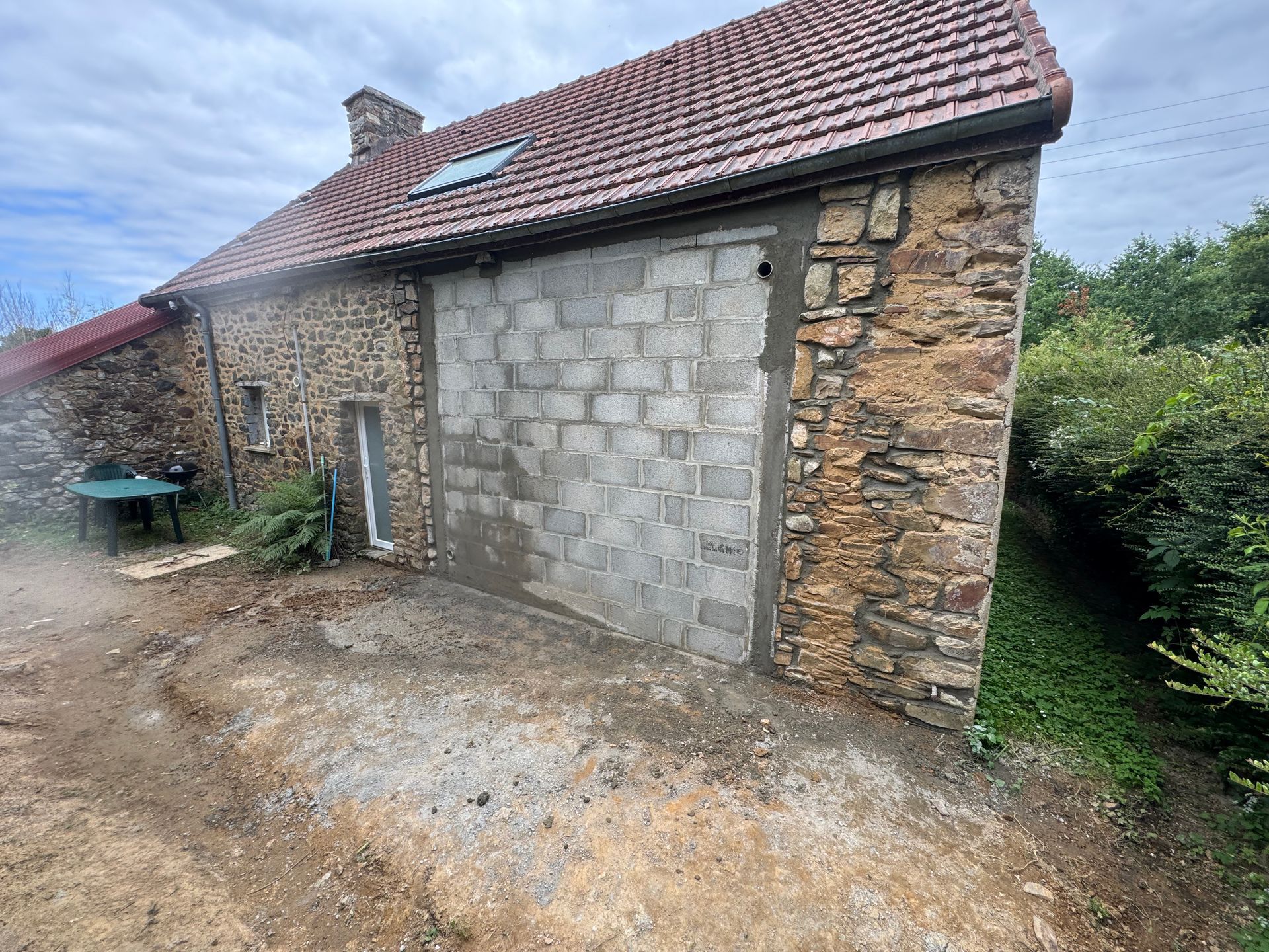 Dépendance en pierre avec toit de tuiles rouges et grande porte de garage grise, à côté d'un chemin de terre et de verdure.