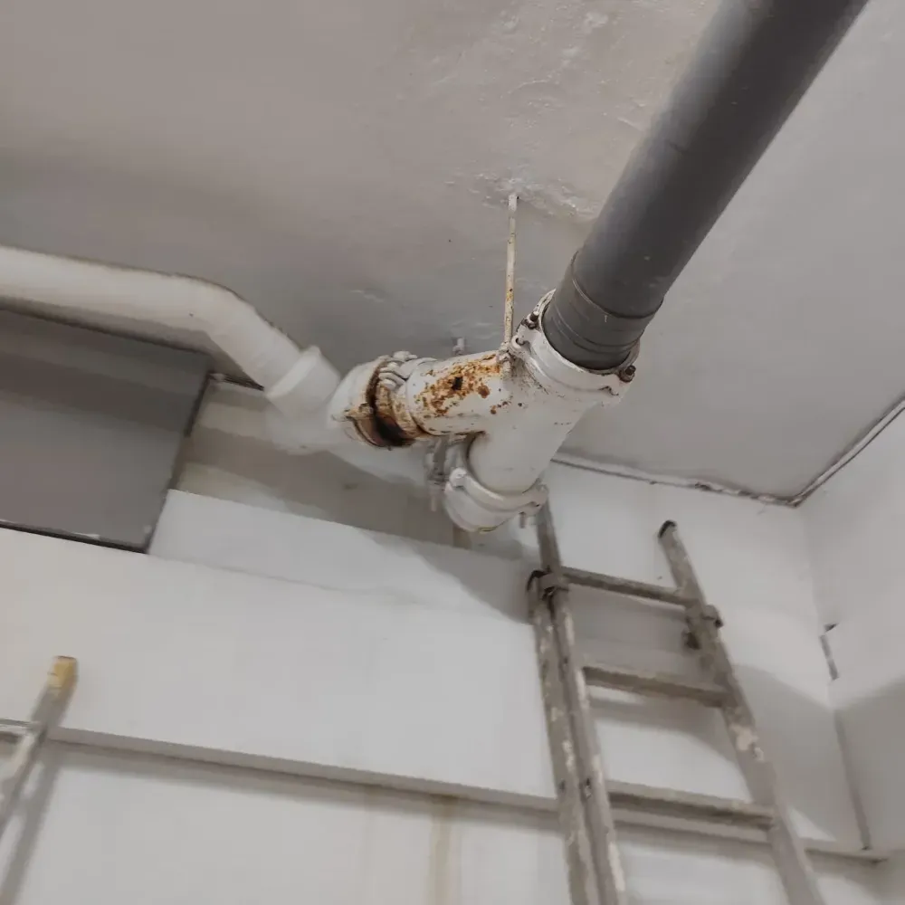 Junta de tubería oxidada cerca del techo con una escalera debajo en un área de servicio interior.