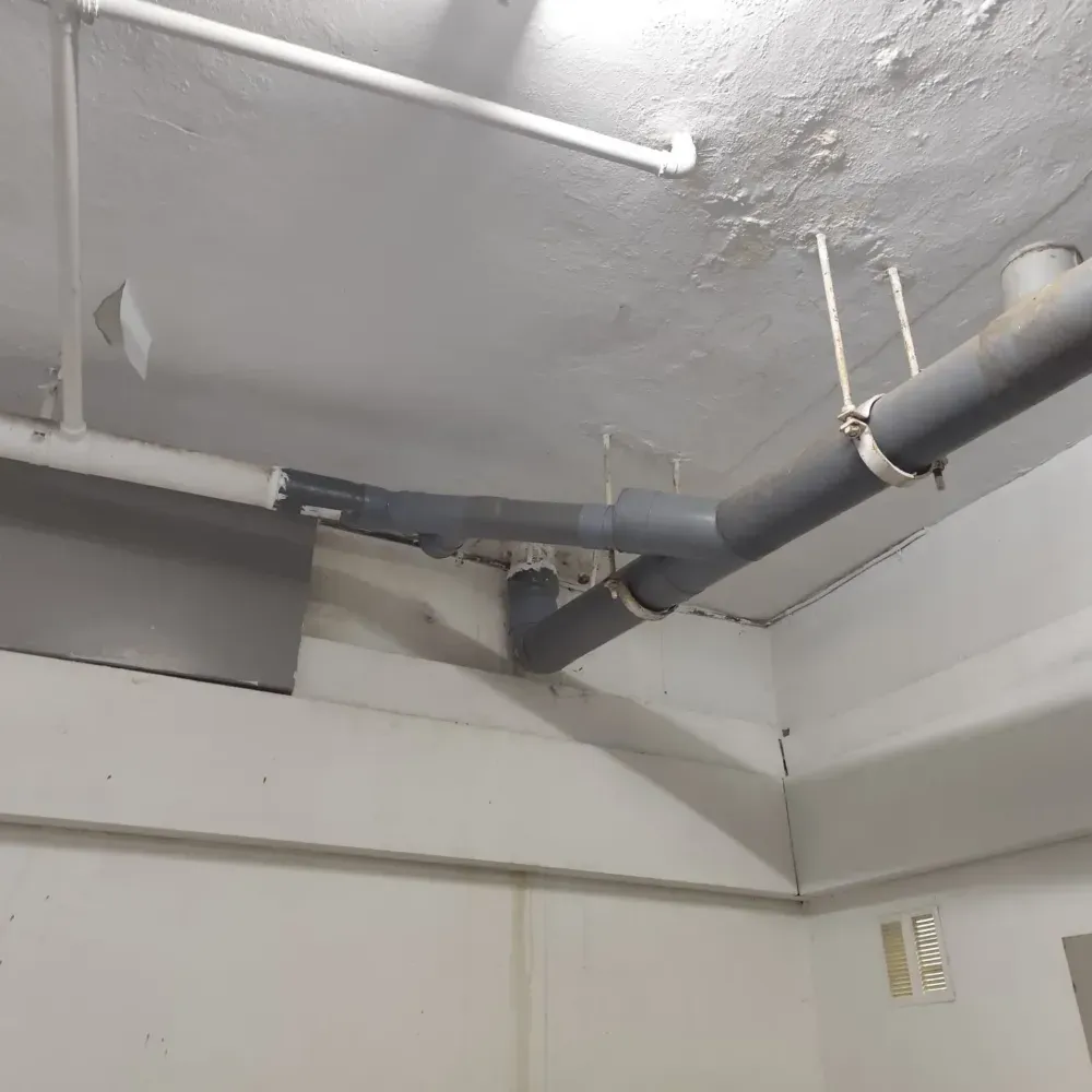 Techo con tuberías grises a la vista, luz fluorescente blanca y pintura descascarada en un cuarto de servicio.
