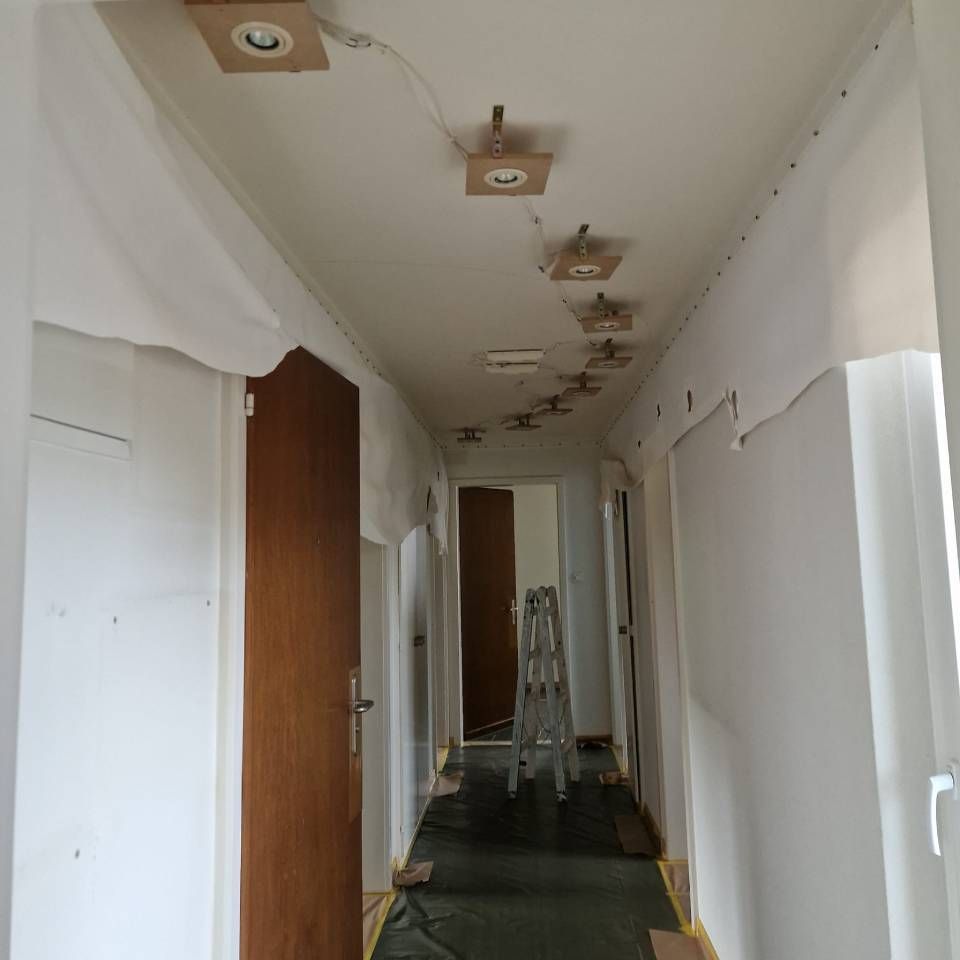 Schmaler, weisser Flur im Bau mit freiliegenden Deckenleuchten, Türen und einer Leiter am anderen Ende