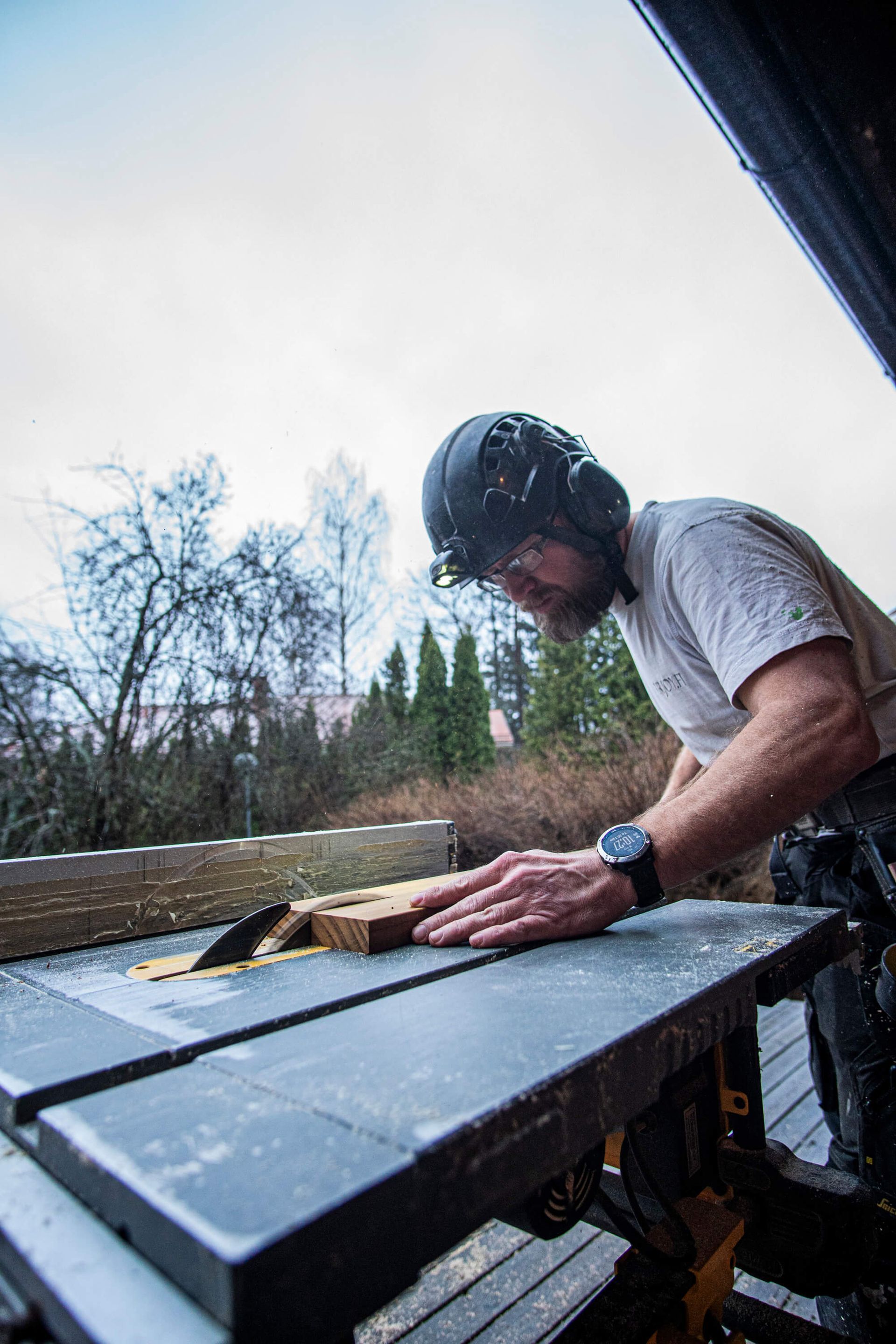 Henkilö käyttää pöytäsahaa ulkona talon vieressä sahaamassa puuta työpöydällä.