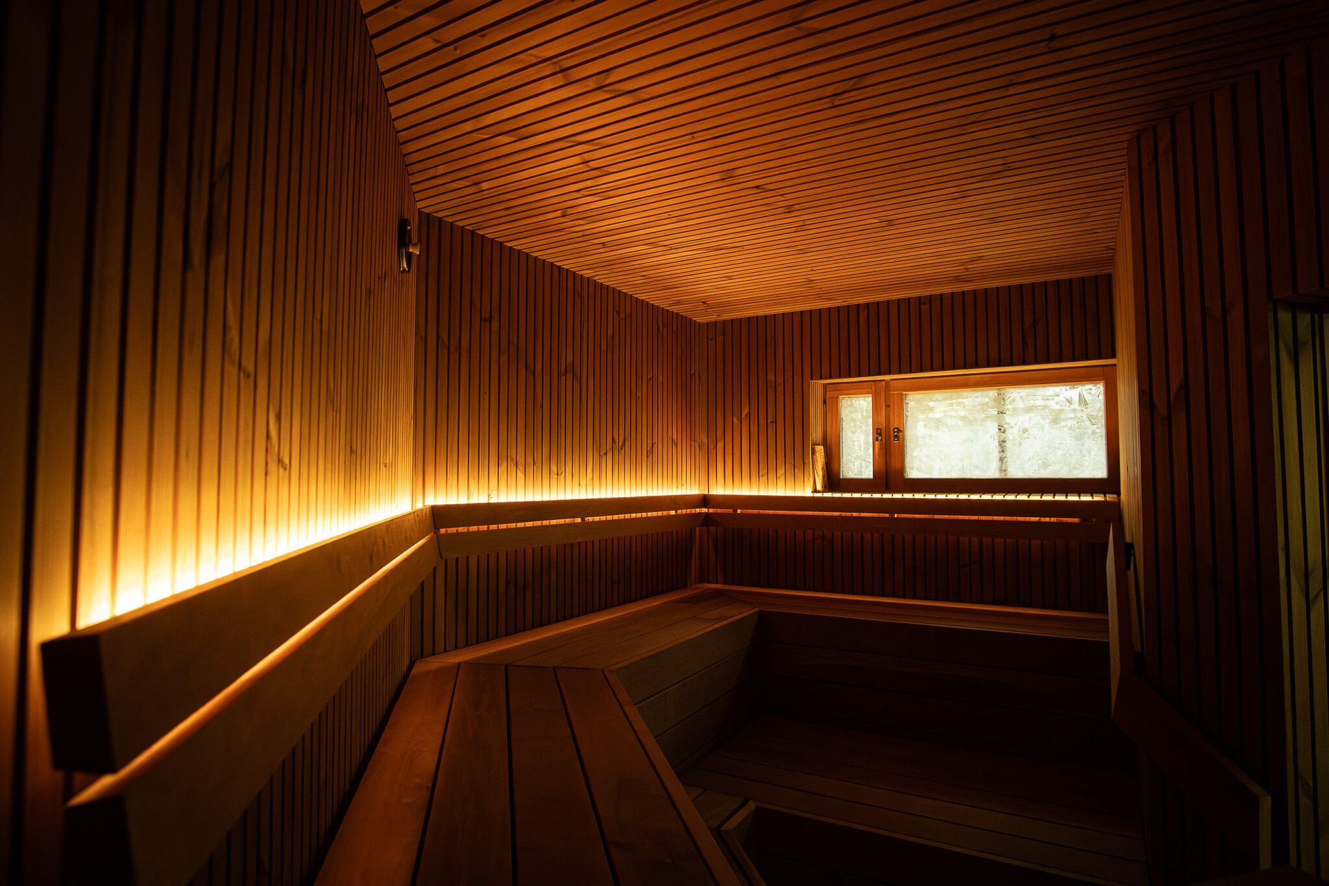 Puupaneloitu sauna, jossa on lämmin valaistus, puiset penkit ja pieni ikkuna.