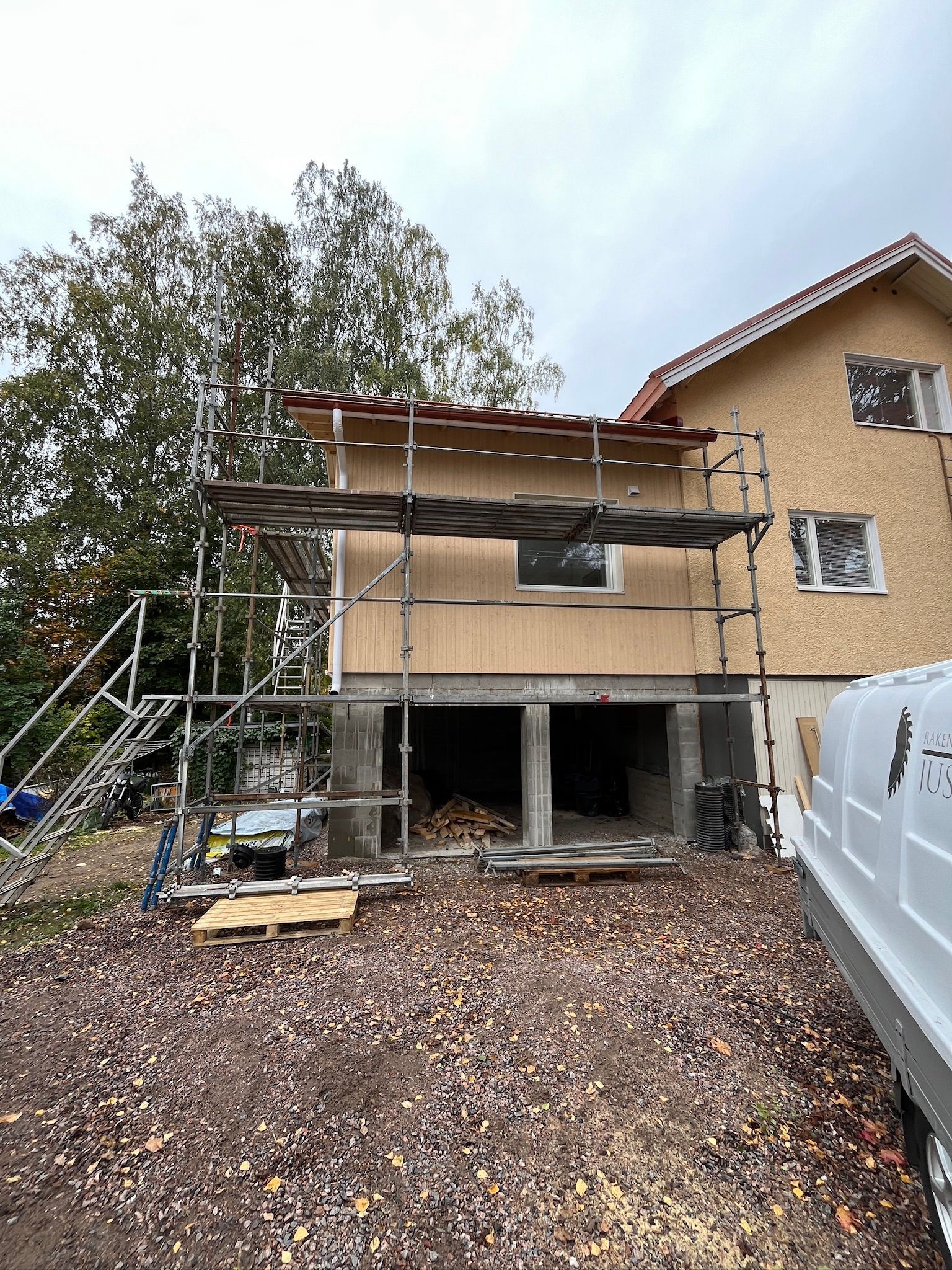Talon julkisivu remontissa, rakennustelineet, soratie ja valkoinen pakettiauto sen vieressä