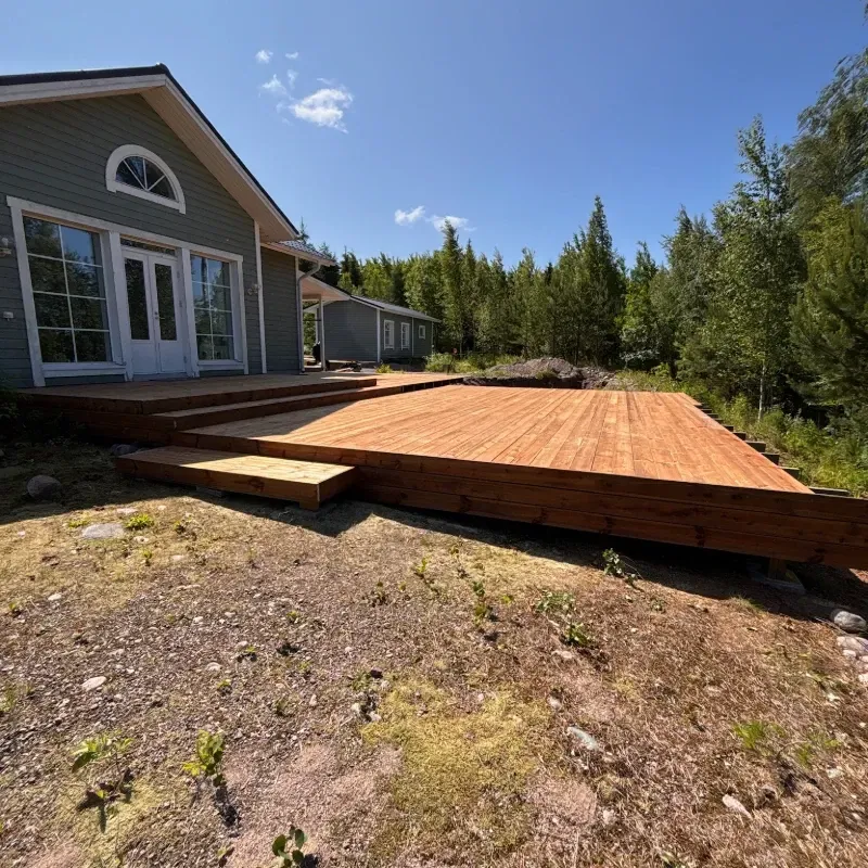 Puinen terassi rakenteilla talon vieressä, portaat ja ympäröivät puut sinisen taivaan alla