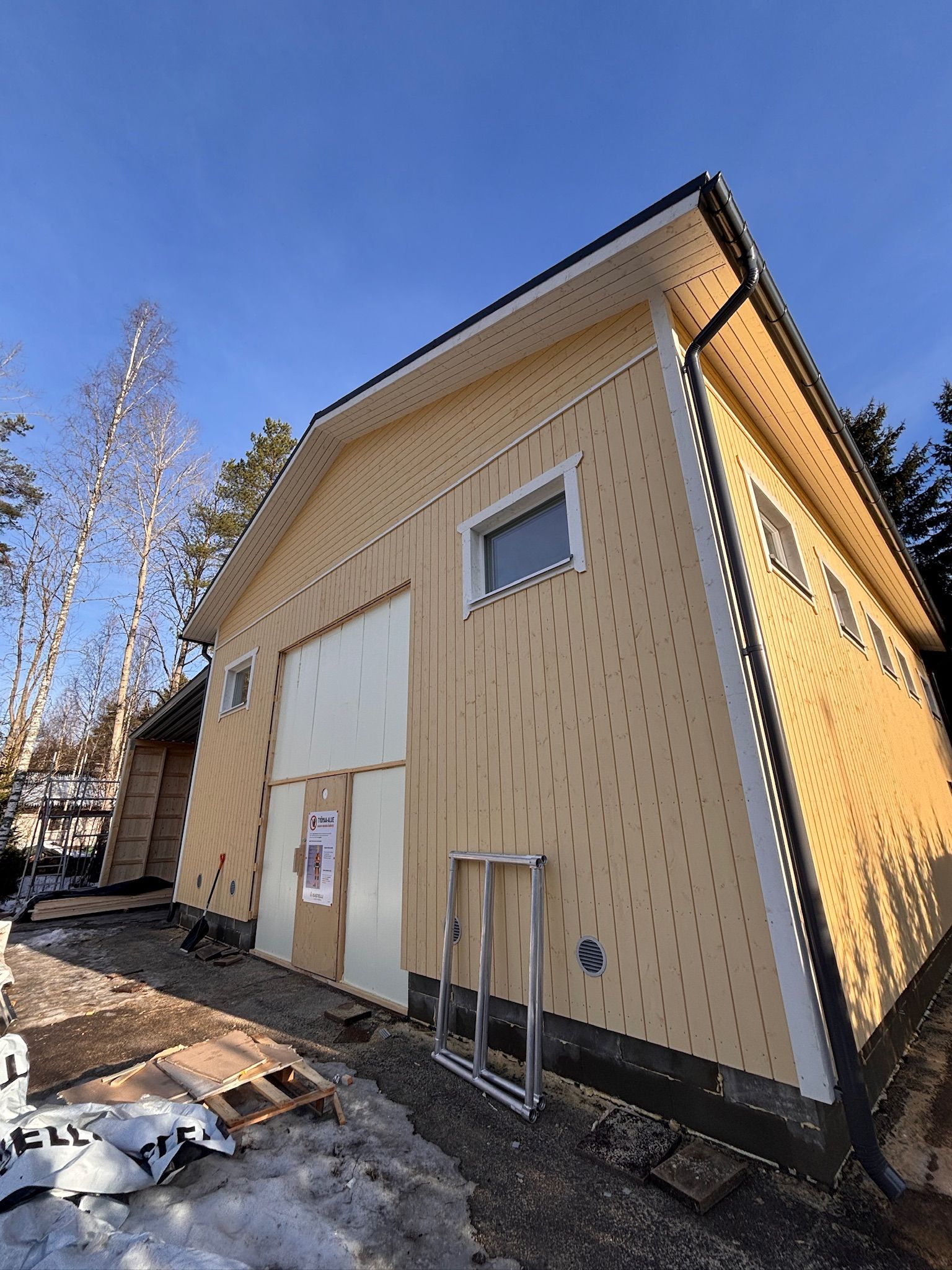 Pieni beigenvärinen talo, jossa on valkoiset ovet ja ikkunat, matalasta kulmasta katsottuna aurinkoisena päivänä.
