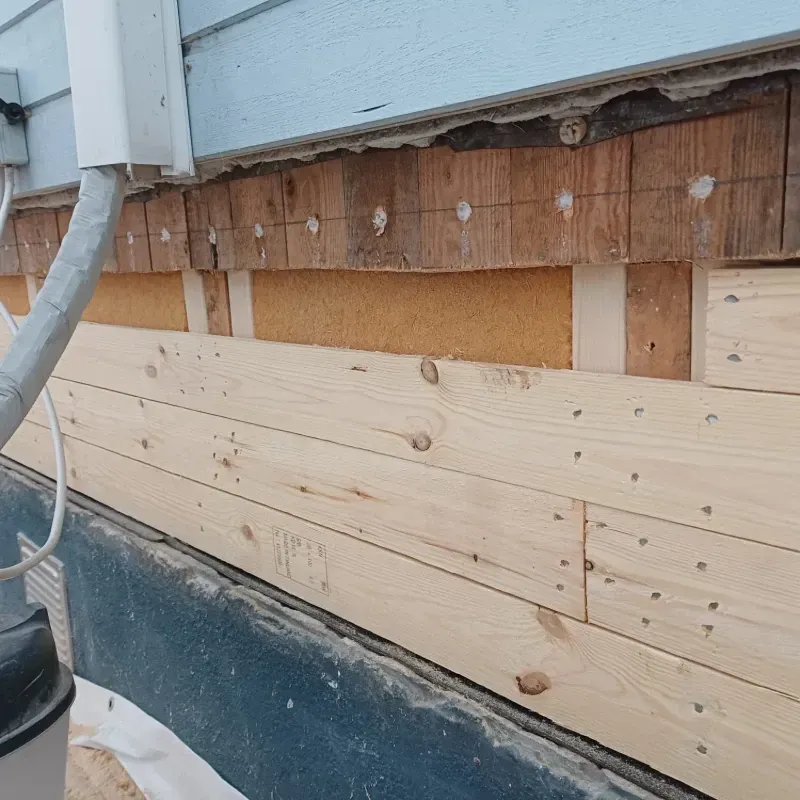 Ulkoseinä, jossa näkyy ulkoverhous ja korjattu puuverhous sinisen räystään alla, syöksytorven vieressä.