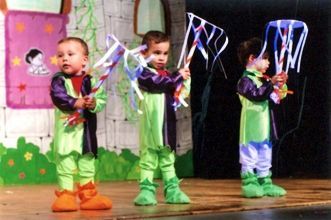 Tres niños vestidos de verde y morado sostienen cometas en un escenario.