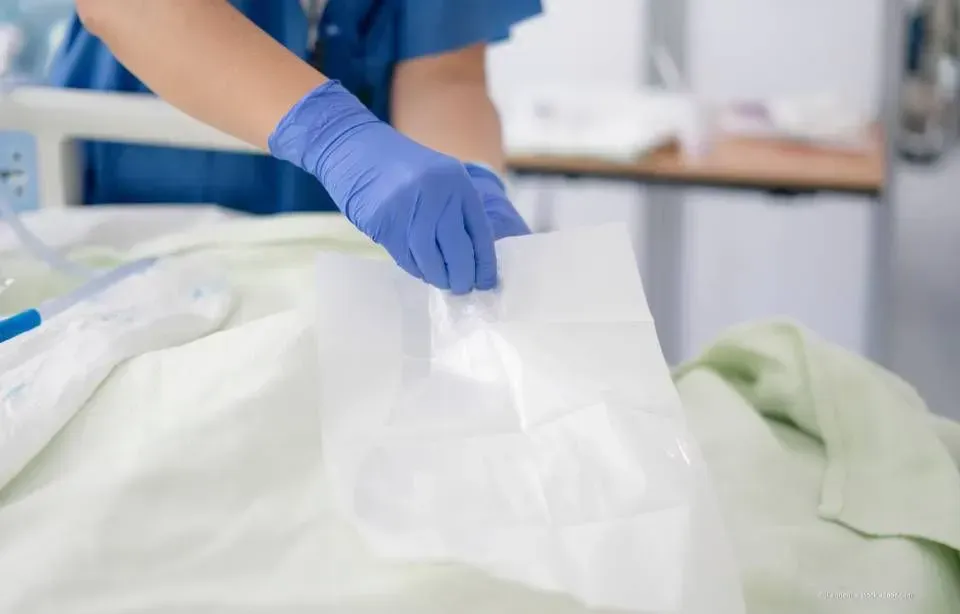 Behandschuhte Hände richten ein weißes Laken auf einem Bett in einer klinischen Umgebung.