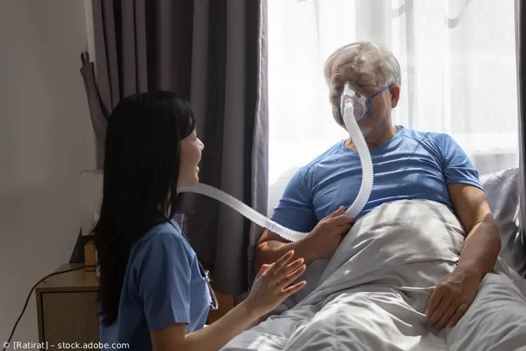 Ein Patient liegt im Krankenhausbett und erhält Sauerstoff, während eine Pflegekraft neben ihm sitzt und seinen Puls prüft.