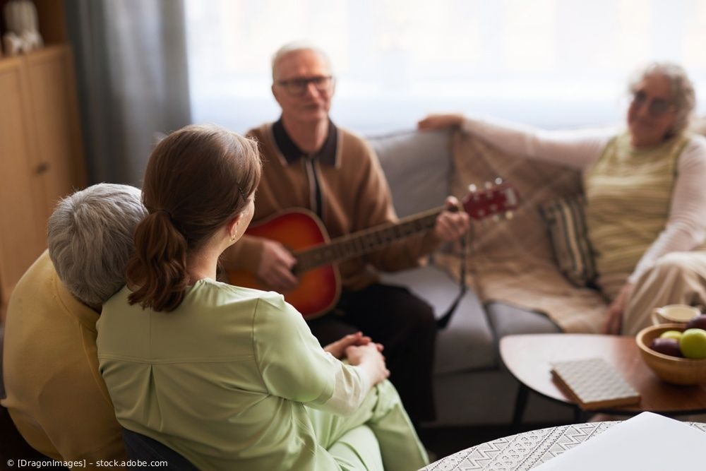 Menschen sitzen in einem Wohnzimmer, einer spielt Gitarre und die anderen hören zu.