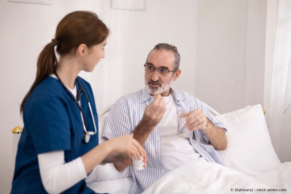 Eine Krankenschwester unterhält sich mit einem Patienten im Bett, der ein Hörgerät hält, in einem hellen Krankenzimmer.