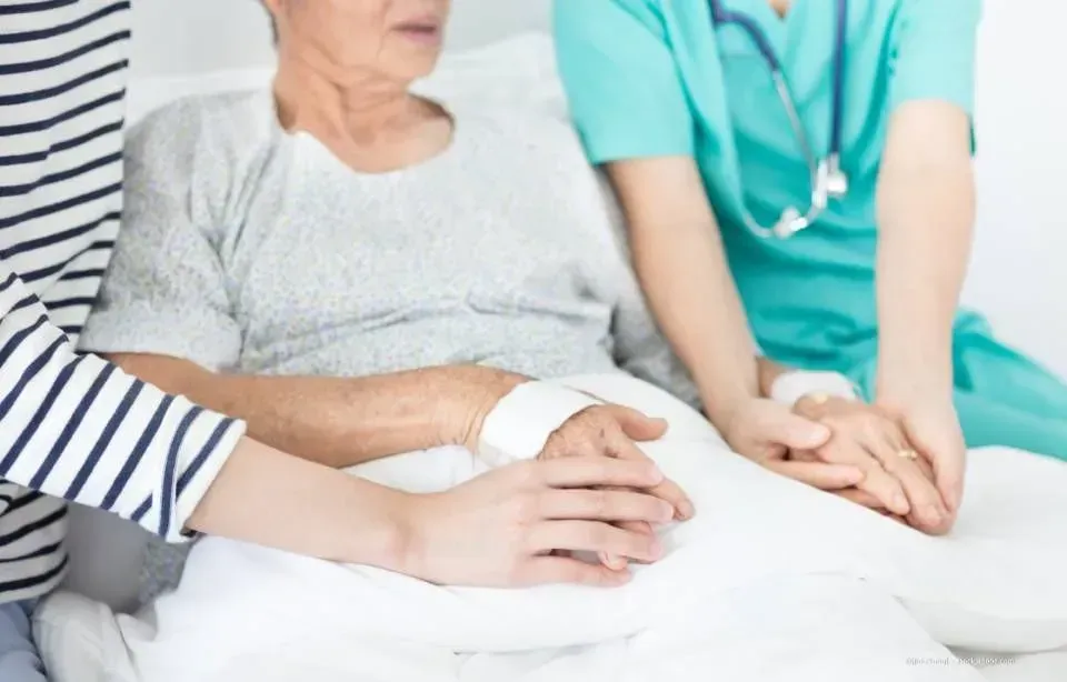 Medizinisches Personal tröstet einen Patienten im Bett, dessen Hände auf der Decke ruhen, in einem hellen Krankenzimmer.