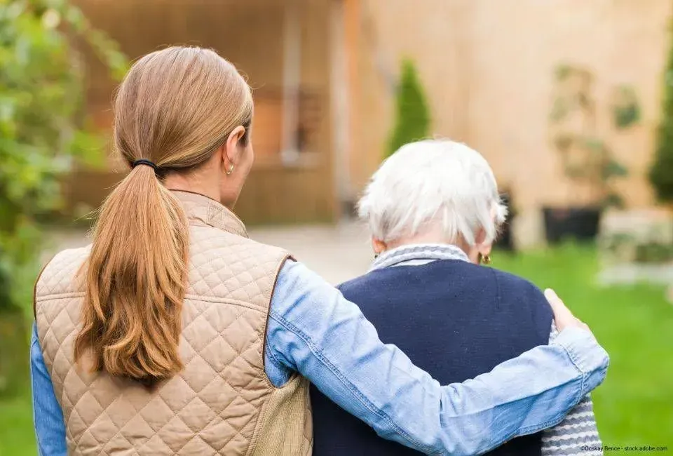 Eine Frau mit Pferdeschwanz geht Arm in Arm mit einer älteren Person auf einem Weg im Freien.