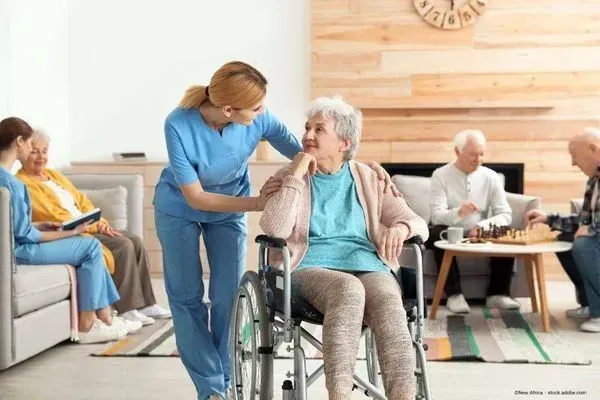 Eine Krankenschwester hilft einer älteren Frau im Rollstuhl in einem hellen Aufenthaltsraum einer Pflegeeinrichtung.