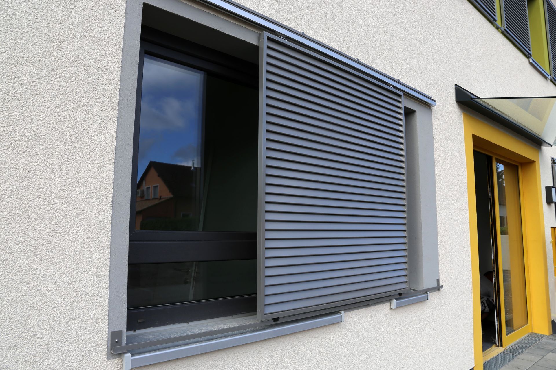 Un volet gris recouvre une fenêtre d'un bâtiment en stuc, à côté d'une porte jaune.