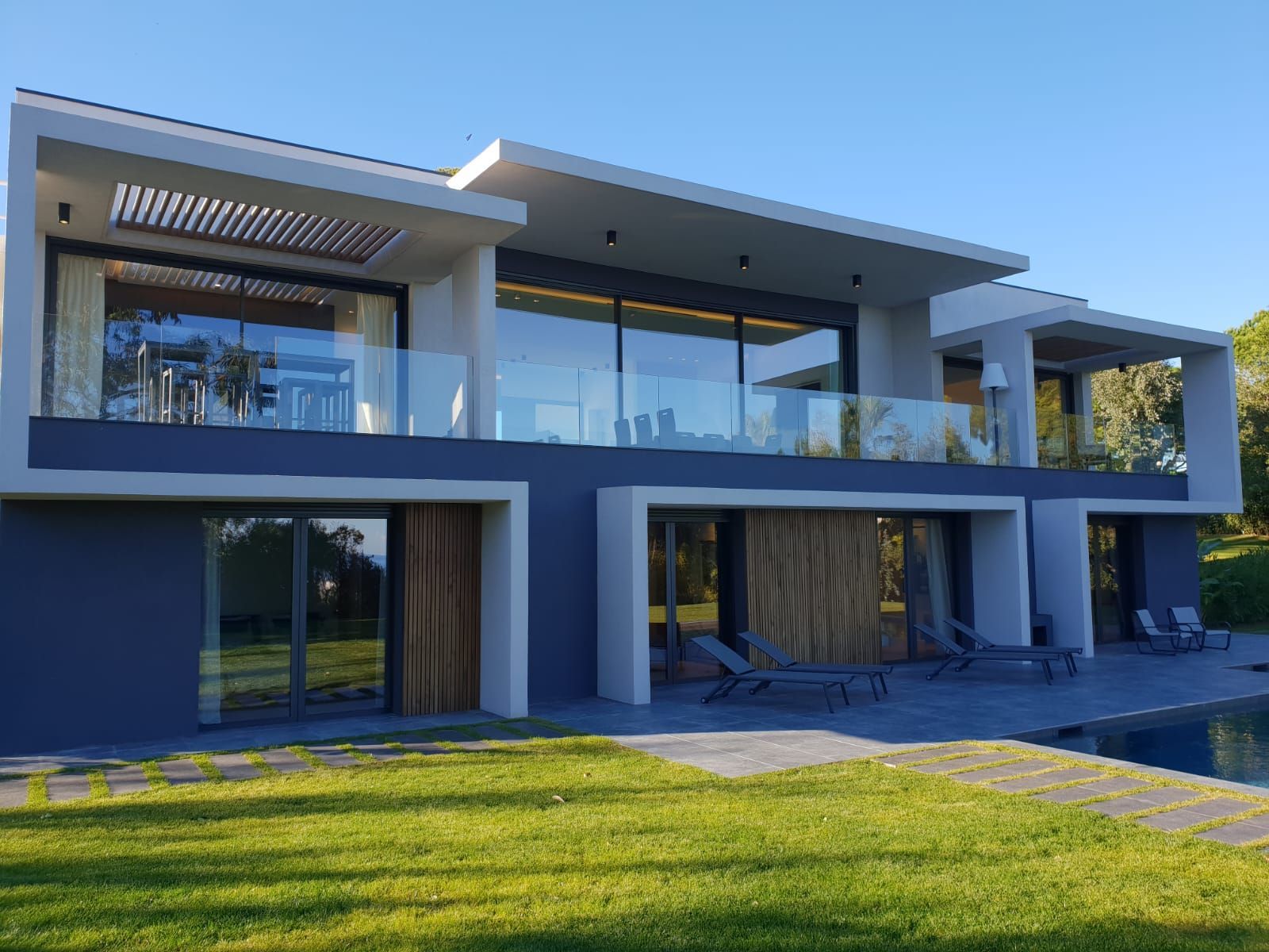 Maison blanche moderne de deux étages avec de grandes baies vitrées, un balcon et une pelouse verte à l'avant.