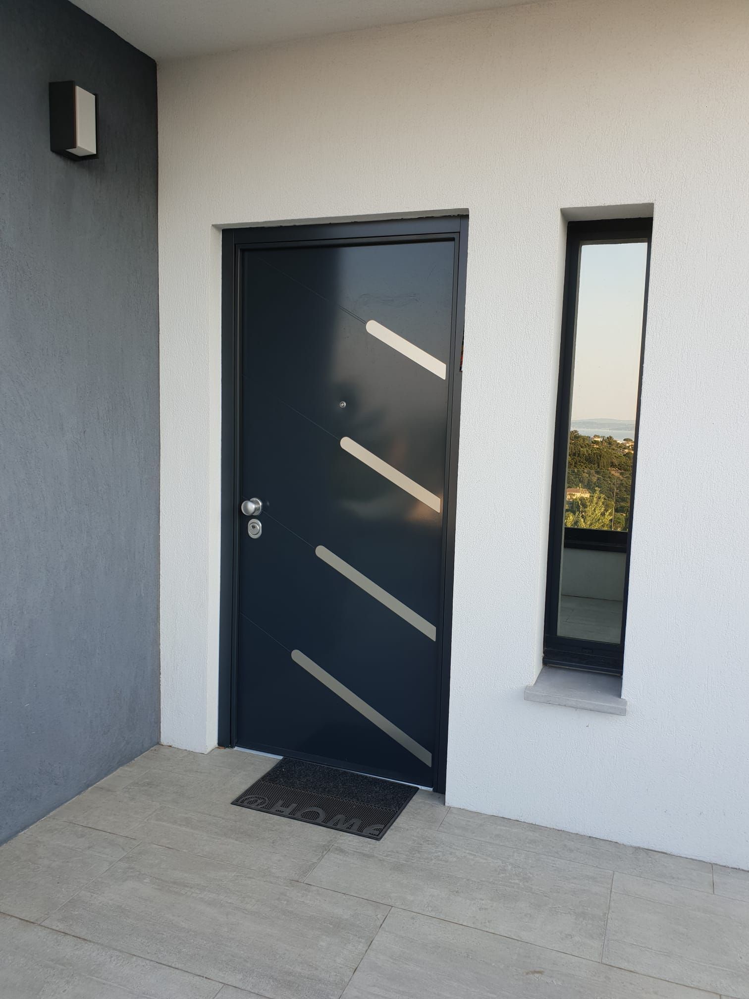 Porte d'entrée moderne avec panneaux de verre diagonaux à côté d'une étroite fenêtre verticale sur un mur extérieur gris