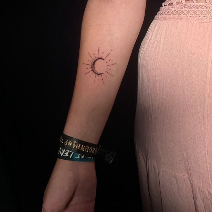 Un tatuaje de una luna creciente con líneas radiantes en el antebrazo de una persona, junto a una prenda rosa.