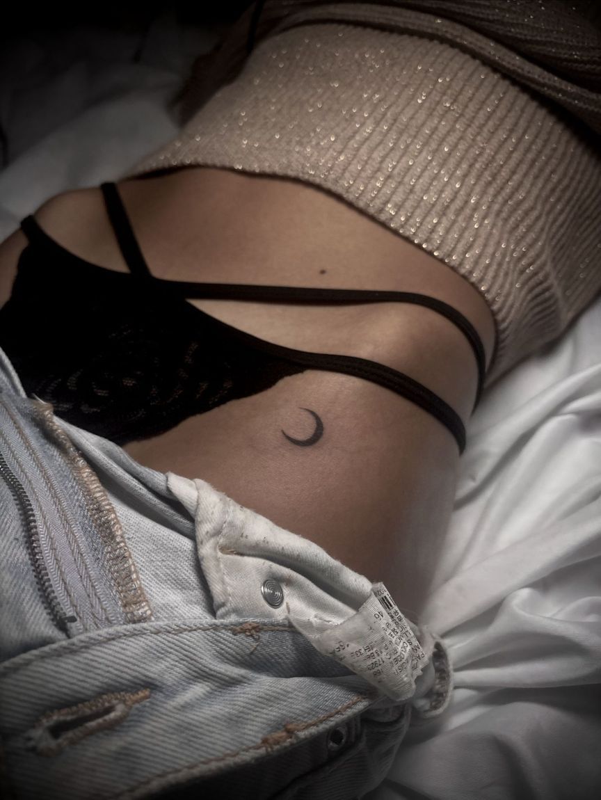 Un tatuaje de una luna creciente en la cadera, con tirantes de lencería oscura visibles sobre la piel