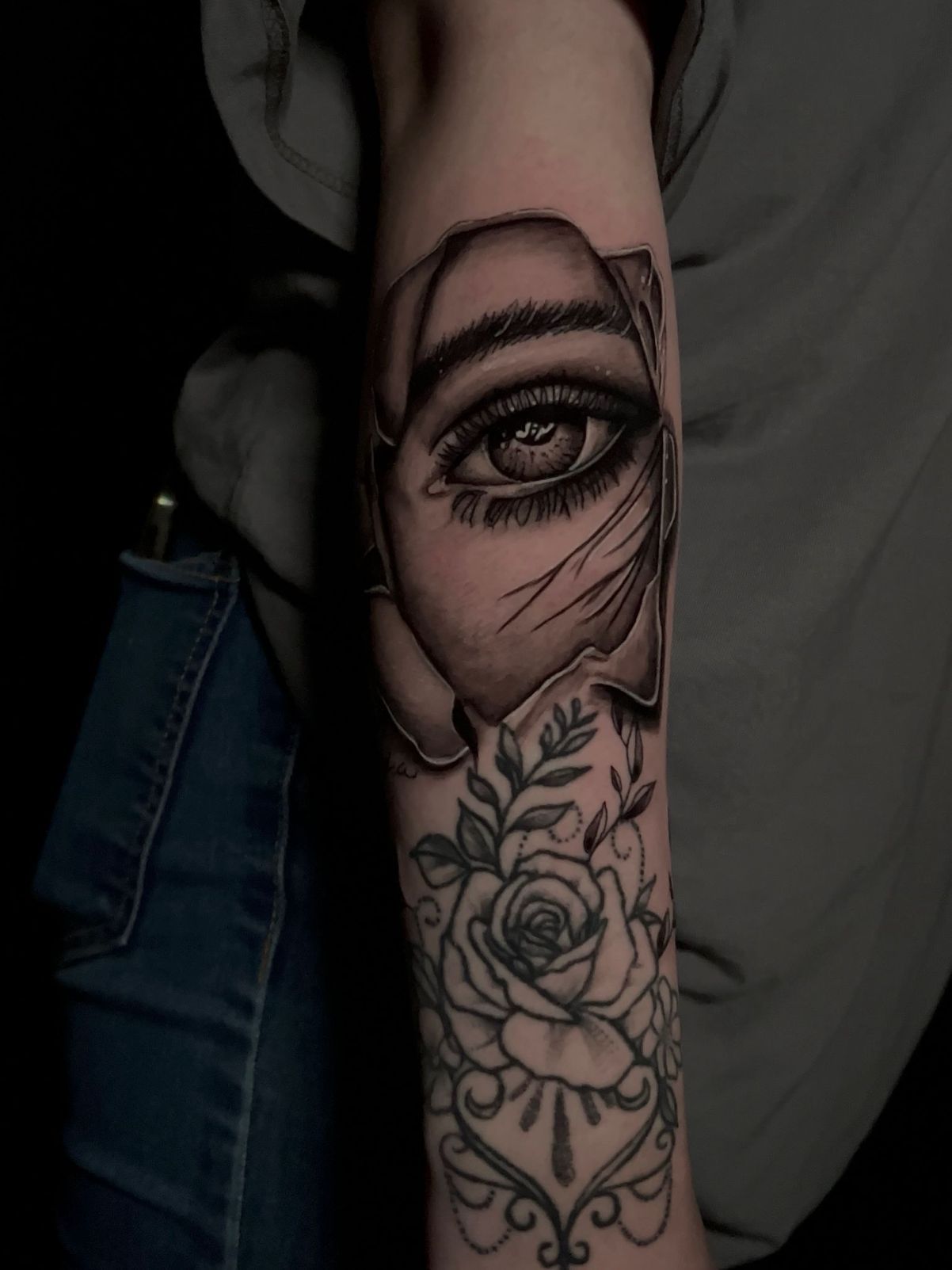 Un tatuaje en el antebrazo que presenta un ojo realista y detallado sobre un diseño floral con rosas y líneas ornamentales.