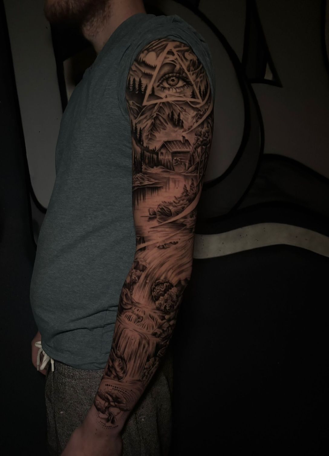 Una persona que muestra un tatuaje en el brazo, hecho de tinta negra y gris, que representa un paisaje, un bosque y un ojo