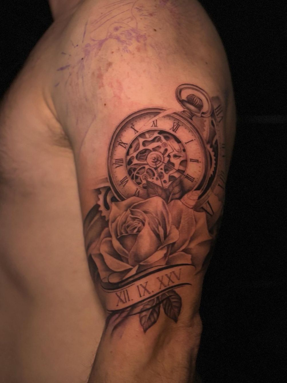 Un tatuaje en blanco y negro en la parte superior del brazo que muestra un reloj de bolsillo, una rosa y números romanos.