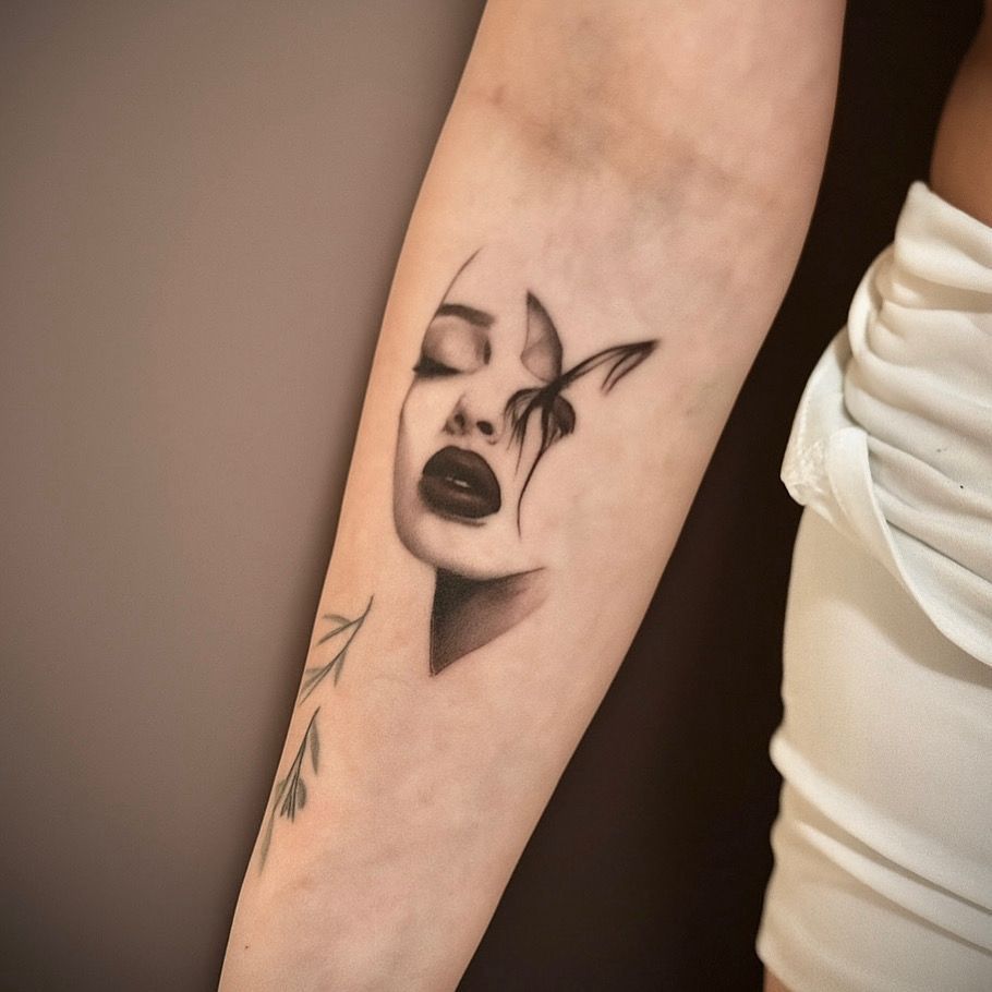 Un tatuaje en blanco y negro en el antebrazo que representa un rostro  en la forma de una mariposa.