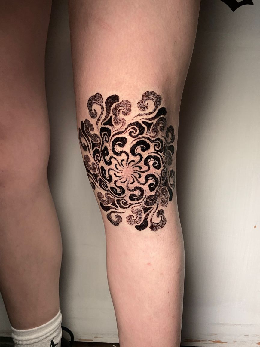 Un diseño de tatuaje ornamental circular, negro y con forma de espiral, centrado en la rodilla de una persona.