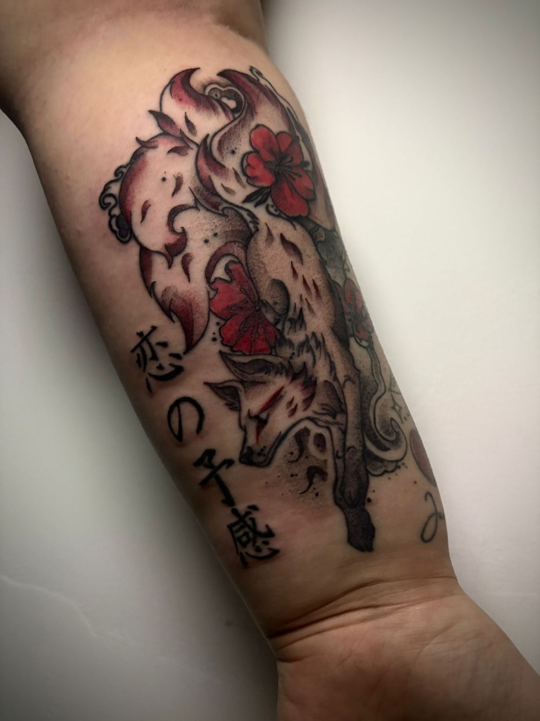 Un tatuaje en el antebrazo con un zorro estilizado de varias colas, flores rojas y texto en caligrafía japonesa.