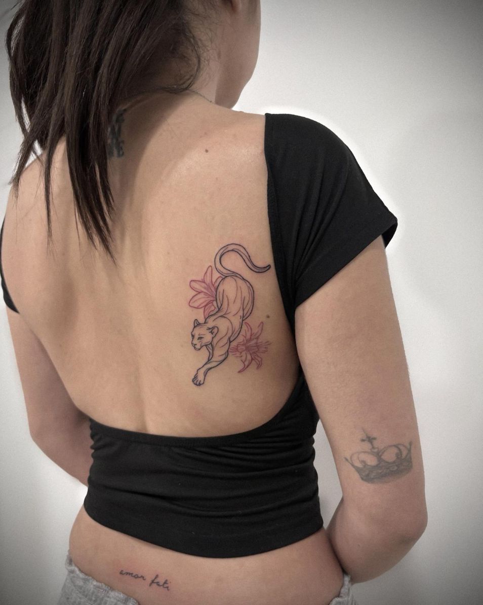 Una persona vista de espaldas, con una camiseta negra, luce un tatuaje lineal de una pantera saltando con flores rosas.